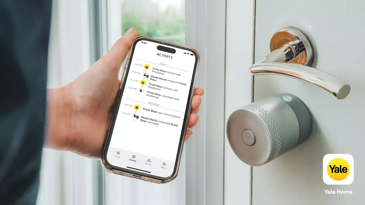 En hand håller en telefon med en Yale Doorman Bluetooth-app som visar aktivitetslogg. Dörren har ett modernt Yale-lås.