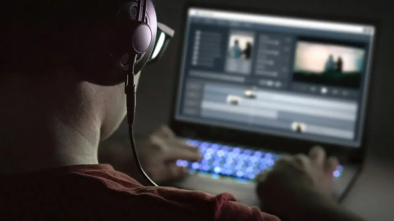 Person med hörlurar och glasögon arbetar med videoredigering på en Mac-dator i mörker. Tangentbordet lyser blått.