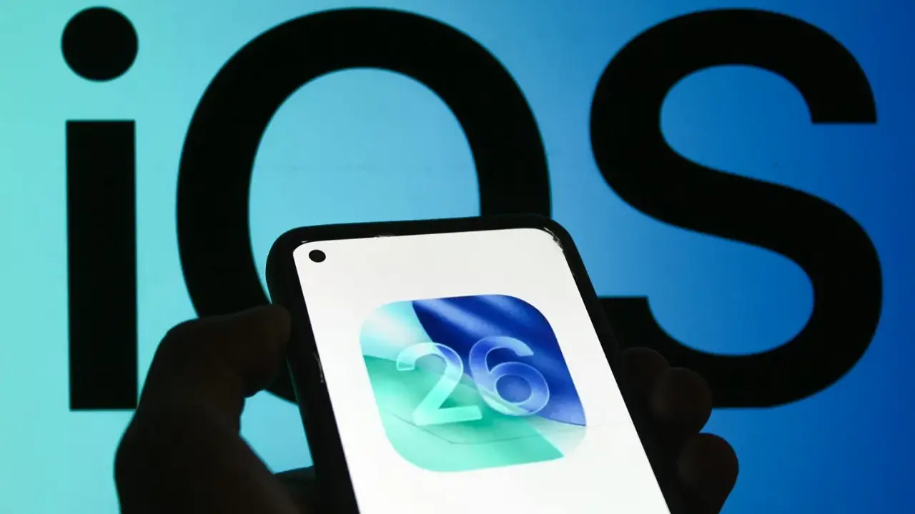 En hand håller en iPhone med ikonen för iOS 26. Bakgrunden visar en stor, suddig logotyp för iOS.