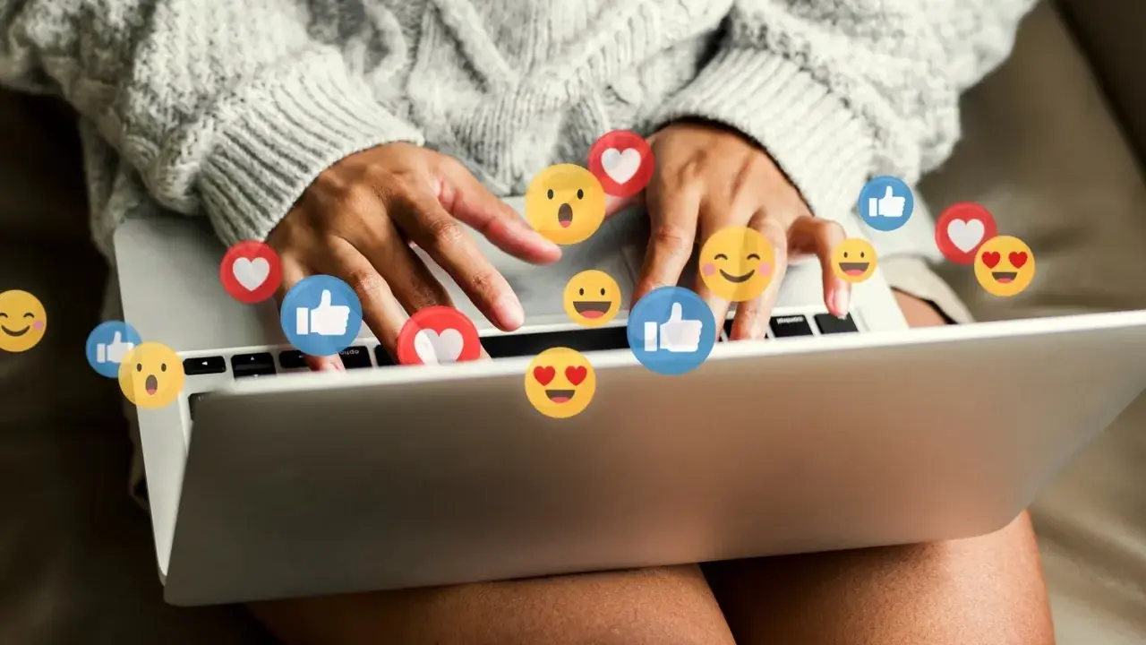 Person skriver på en laptop, omgiven av flygande emojis som hjärtan och tummen upp. Ett kortkommando emoji för att uttrycka känslor.