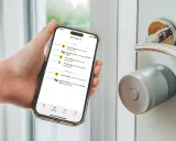 En hand håller en telefon med en Yale Doorman Bluetooth-app som visar aktivitetslogg. Dörren har ett modernt Yale-lås.