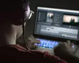 Person med hörlurar och glasögon arbetar med videoredigering på en Mac-dator i mörker. Tangentbordet lyser blått.