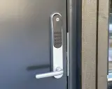 Yale doorman blinkar grönt. Dörren är grå och har ett modernt handtag. Bredvid syns en träfasad och ett fönster med snö utanför.