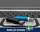 Två USB-minnen, ett svart och ett blått, ligger på ett tangentbord. En guide för Windows 11 USB på svenska.