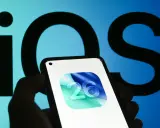 En hand håller en iPhone med ikonen för iOS 26. Bakgrunden visar en stor, suddig logotyp för iOS.