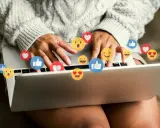 Person skriver på en laptop, omgiven av flygande emojis som hjärtan och tummen upp. Ett kortkommando emoji för att uttrycka känslor.