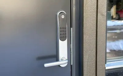 Yale doorman blinkar grönt. Dörren är grå och har ett modernt handtag. Bredvid syns en träfasad och ett fönster med snö utanför.