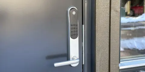 Yale doorman blinkar grönt. Dörren är grå och har ett modernt handtag. Bredvid syns en träfasad och ett fönster med snö utanför.