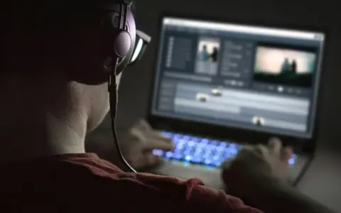 Person med hörlurar och glasögon arbetar med videoredigering på en Mac-dator i mörker. Tangentbordet lyser blått.