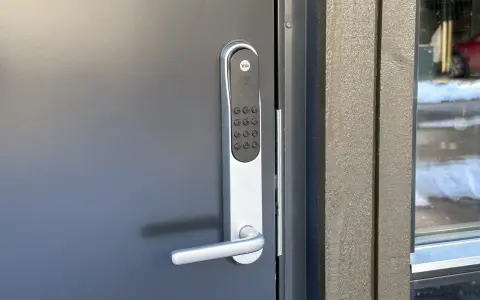 Yale doorman blinkar grönt. Dörren är grå och har ett modernt handtag. Bredvid syns en träfasad och ett fönster med snö utanför.