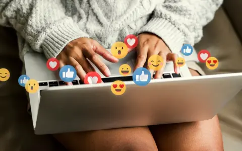 Person skriver på en laptop, omgiven av flygande emojis som hjärtan och tummen upp. Ett kortkommando emoji för att uttrycka känslor.