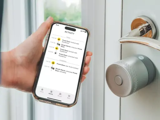En hand håller en telefon med en Yale Doorman Bluetooth-app som visar aktivitetslogg. Dörren har ett modernt Yale-lås.