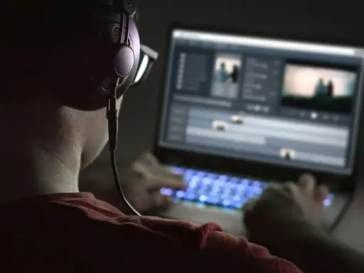 Person med hörlurar och glasögon arbetar med videoredigering på en Mac-dator i mörker. Tangentbordet lyser blått.