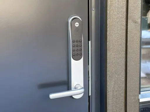 Yale doorman blinkar grönt. Dörren är grå och har ett modernt handtag. Bredvid syns en träfasad och ett fönster med snö utanför.