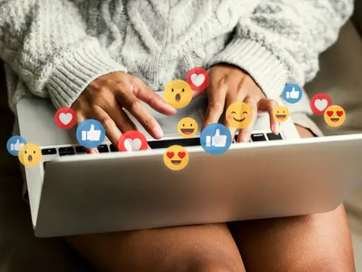 Person skriver på en laptop, omgiven av flygande emojis som hjärtan och tummen upp. Ett kortkommando emoji för att uttrycka känslor.