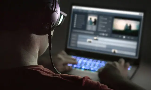 Person med hörlurar och glasögon arbetar med videoredigering på en Mac-dator i mörker. Tangentbordet lyser blått.