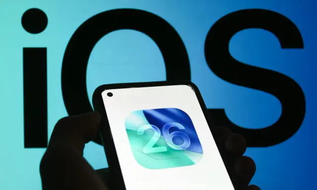 En hand håller en iPhone med ikonen för iOS 26. Bakgrunden visar en stor, suddig logotyp för iOS.