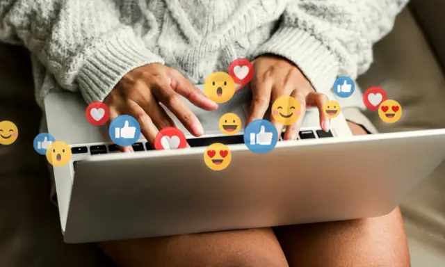 Person skriver på en laptop, omgiven av flygande emojis som hjärtan och tummen upp. Ett kortkommando emoji för att uttrycka känslor.