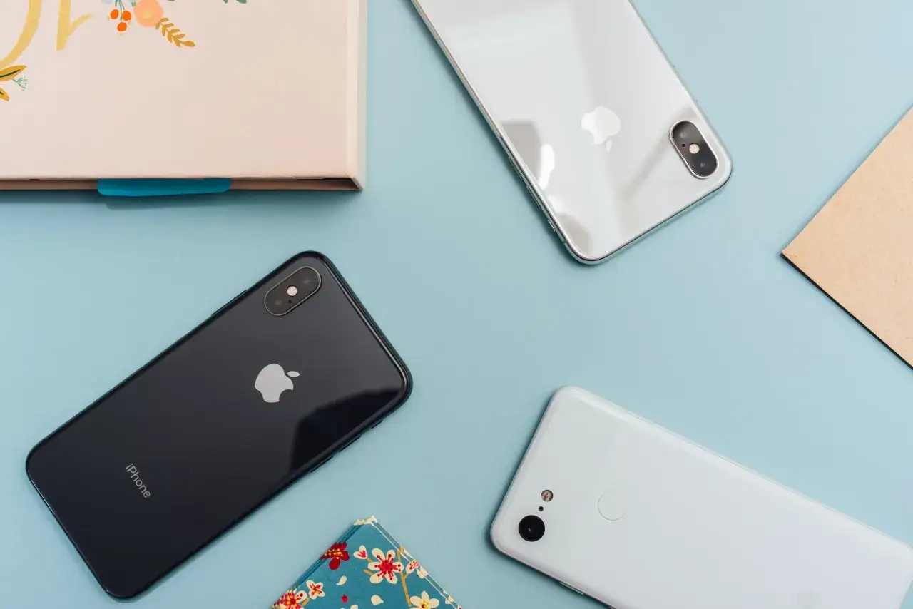 Två iPhones och en Android-telefon ligger på ett ljusblått underlag. En av iPhones är svart, den andra är vit. Android-telefonen är vit.