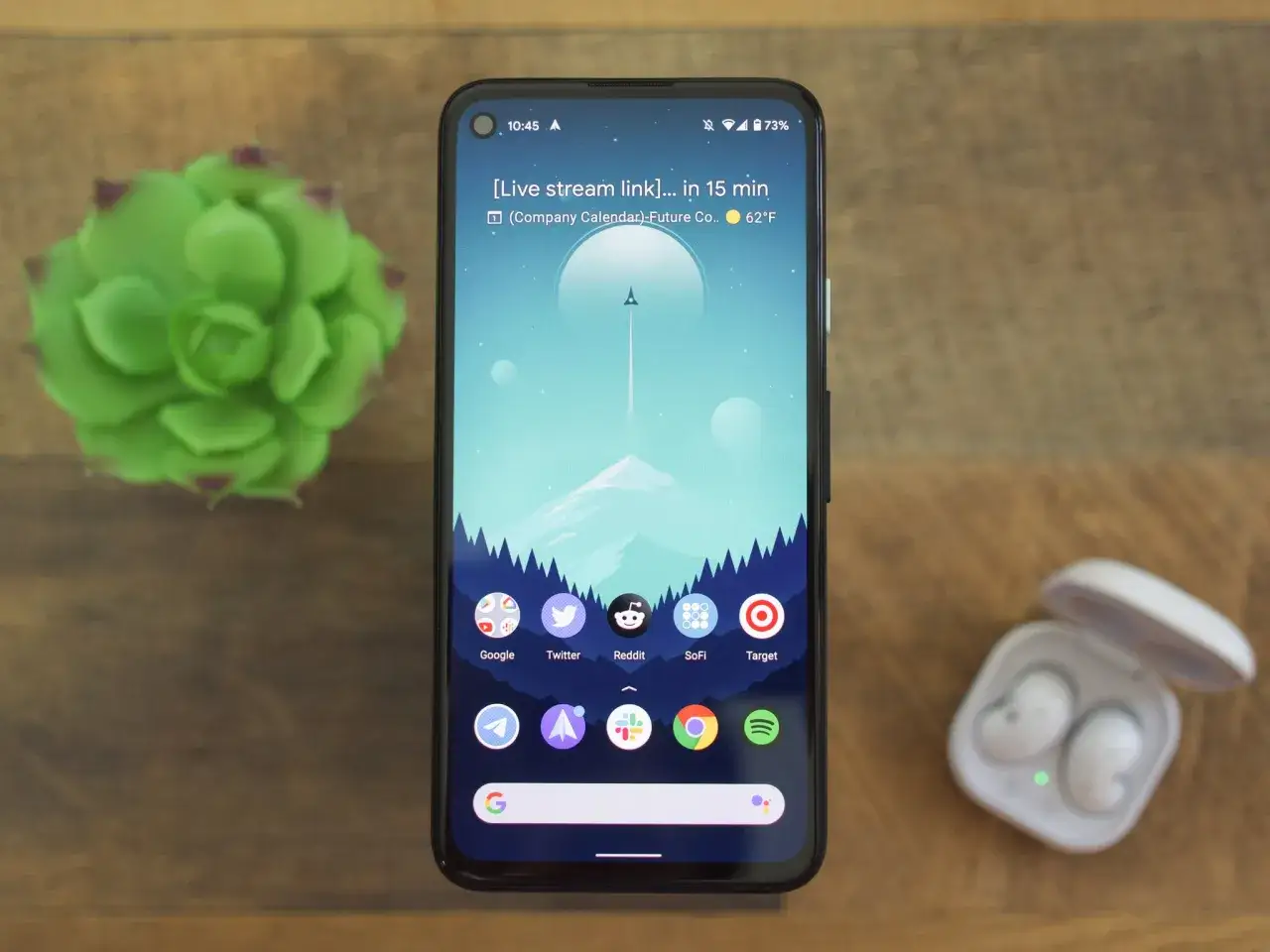 Android-telefon med widgets och appar som Google, Twitter, Reddit, SoFi och Target. En grön suckulent och vita hörlurar ligger bredvid.