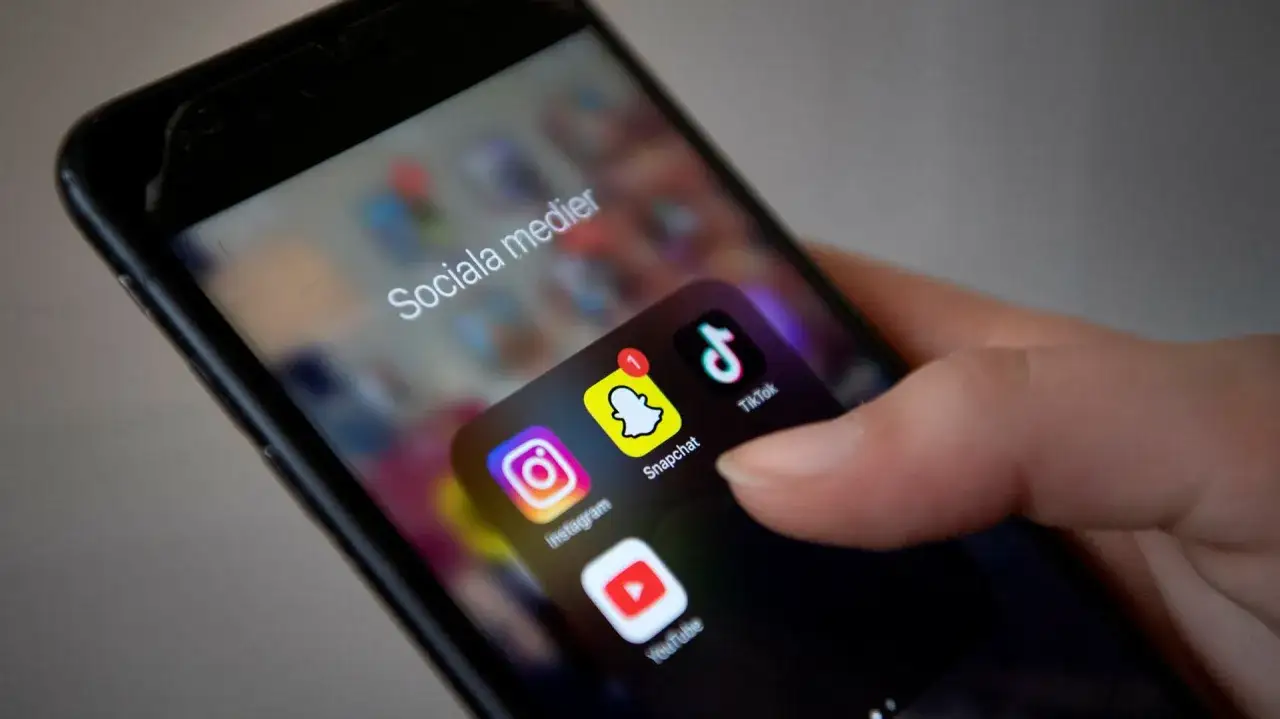 En hand håller en mobiltelefon med ikoner för Instagram, Snapchat, TikTok och YouTube. Texten
