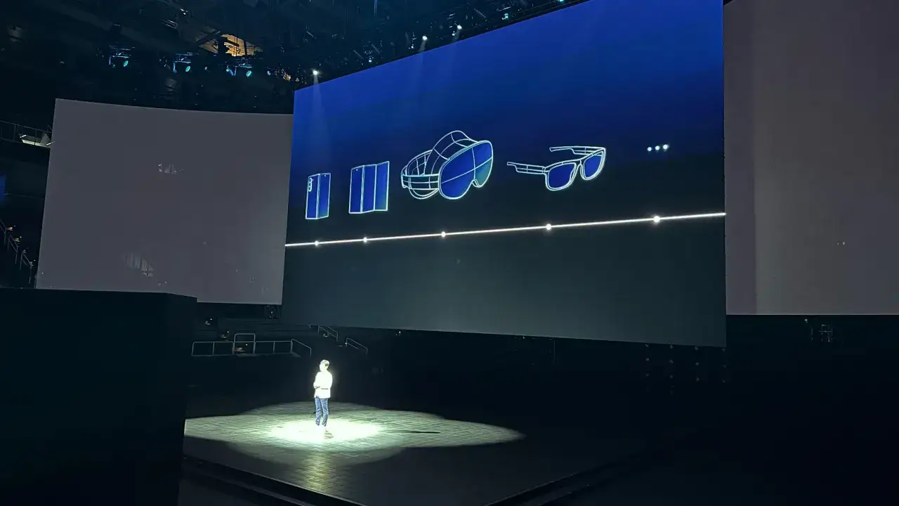 En person står på scenen under en presentation. På den stora skärmen visas en illustration av en vikbar telefon, ett VR-headset och glasögon, som en galaxy unpacked.