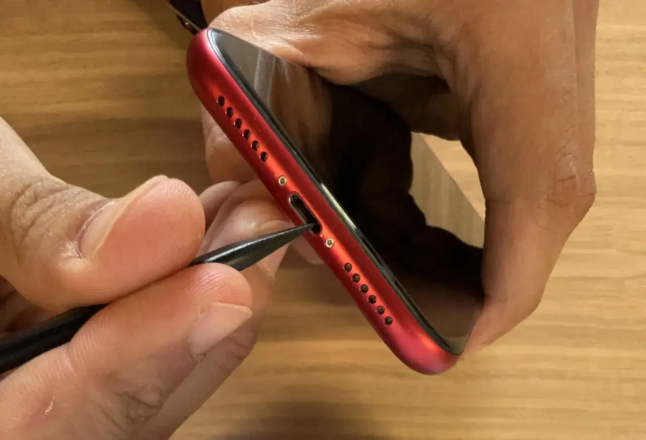 En person f&ouml;rs&ouml;ker reng&ouml;ra laddningsporten p&aring; en r&ouml;d mobiltelefon med ett tunt verktyg, eftersom mobilen laddar inte.
