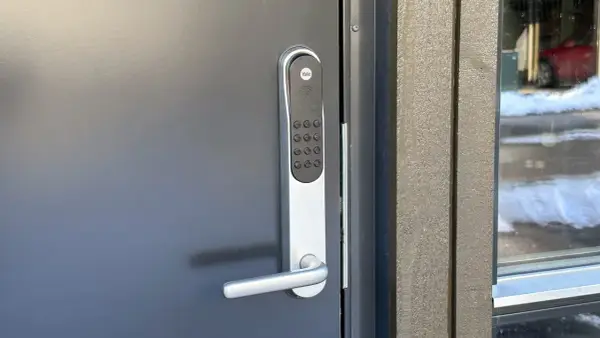 Yale doorman blinkar grönt. Dörren är grå och har ett modernt handtag. Bredvid syns en träfasad och ett fönster med snö utanför.
