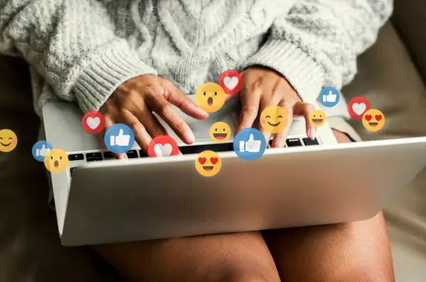 Person skriver på en laptop, omgiven av flygande emojis som hjärtan och tummen upp. Ett kortkommando emoji för att uttrycka känslor.
